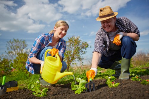 A man and a young woman are working together in a well-maintained garden, planting young green seedlings in rich, dark soil within a neatly edged flower bed. The man, dressed in a checked shirt, blue jeans, and a straw sun hat, is kneeling on the ground and using his hands to carefully position the plants, while the woman, wearing a plaid shirt, blue jeans, and yellow gardening gloves, is holding a yellow watering can, watering the young plants. The garden surrounds them with lush grass, and there are a few trees visible in the background under a bright, partly cloudy sky, indicating a pleasant day suitable for outdoor gardening. The scene is typical of residential garden maintenance, with clean, organized planting beds and a natural outdoor environment, aligning with gardening and lawn care services such as hedge trimming and landscape upkeep in Clapham or nearby areas.