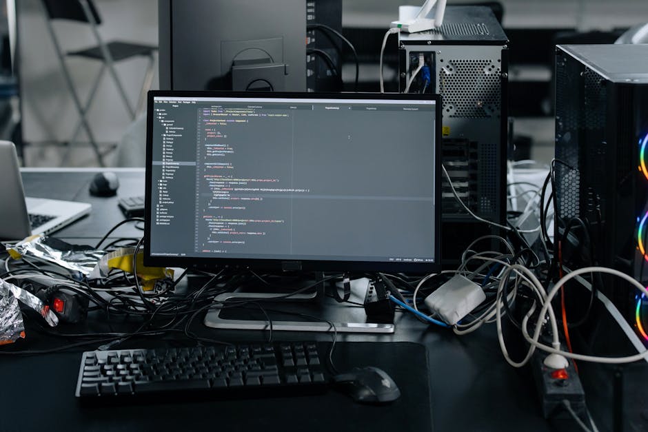A cluttered desk with a computer monitor displaying lines of code, surrounded by tangled cables, a computer mouse, and a keyboard. To the left, there are a laptop and some crumpled foil wrappers. In t